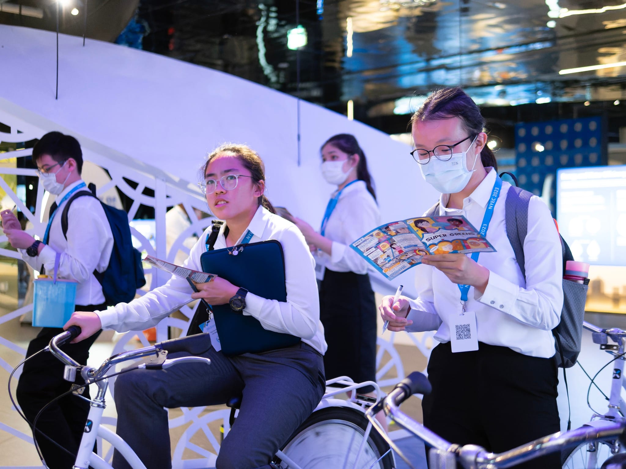 Four young people in white shirts and lanyards at an indoor event, two seated on white bikes reading pamphlets.